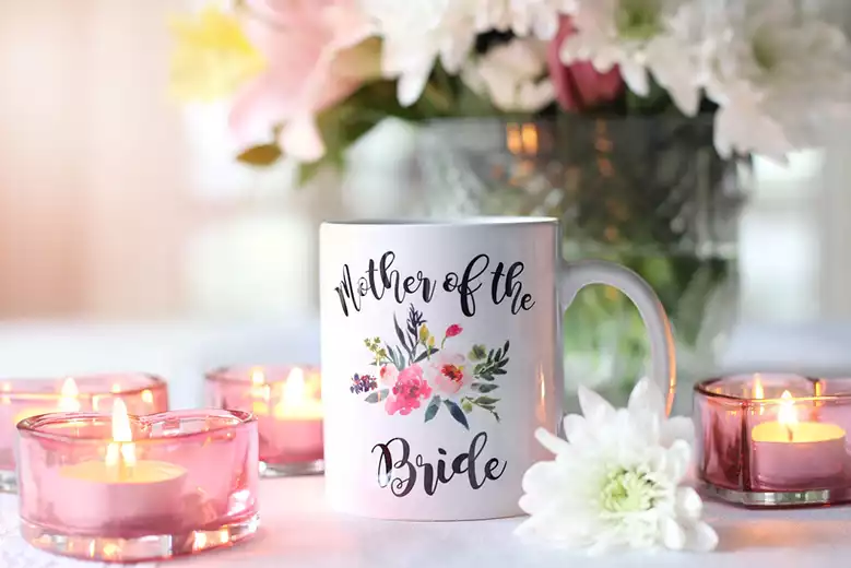 Taza blanca con la leyenda "mother of the bride" sobre mesa rodeada de velas.